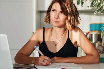 Femme pensive assise devant son carnet, en train d'écrire, qui regarde dans le vide