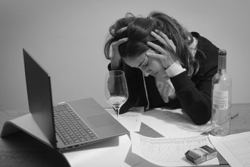 Femme avec un verre d'alcool près de documents et d'un paquet de cigarettes, elle se tient la tête avec les mains et elle a les yeux fermés